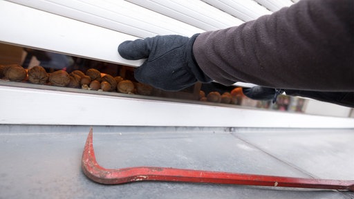 Hände in Handschuhen öffnen mit Gewalt Rollläden. Auf der Fensterbank liegt ein Brecheisen.