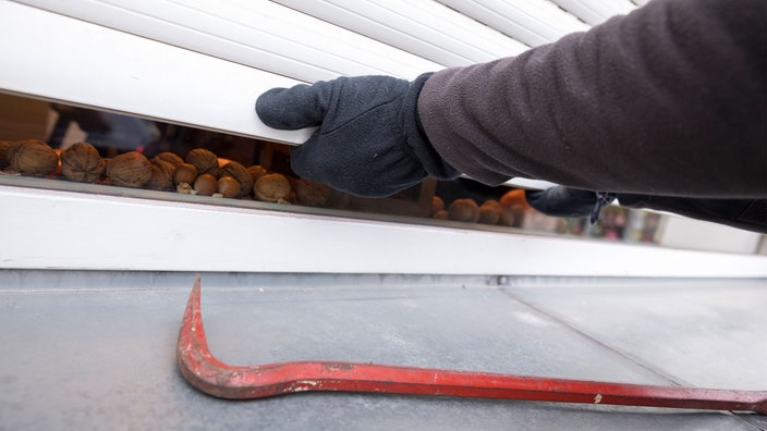 Hände in Handschuhen öffnen mit Gewalt Rollläden. Auf der Fensterbank liegt ein Brecheisen.