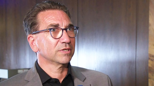 Guido Déus, ein braunhaariger Mann mit Brille im Leinenanzug, spricht.