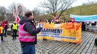 Auf einem Platz haben sich mehrere Menschen mit Bannern für einen Warnstreik versammelt