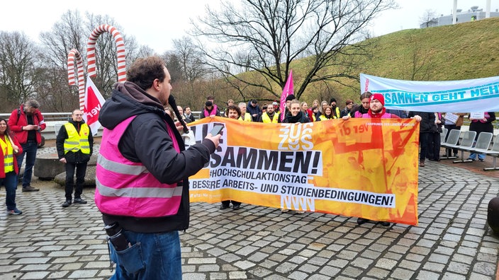 Auf einem Platz haben sich mehrere Menschen mit Bannern für einen Warnstreik versammelt
