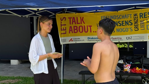 Eine Frau spricht mit einem Jungen an einem Infostand, dahinter die Aufschrift "Let's talk about...".