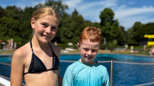 Ein Mädchen und ein Junge stehen an einem Schwimmbecken und lächeln in die Kamera.