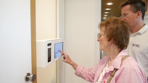 Eine Patientin steht vor einem Tablet und bedient es, rechts von ihr steht Apotheker Oliver Skopp.