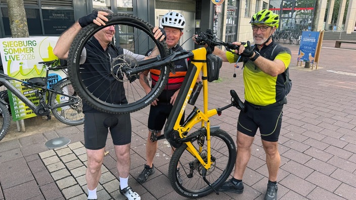 Drei Männer in Fahrradkleidung reparieren ein gelbes Fahrrad.