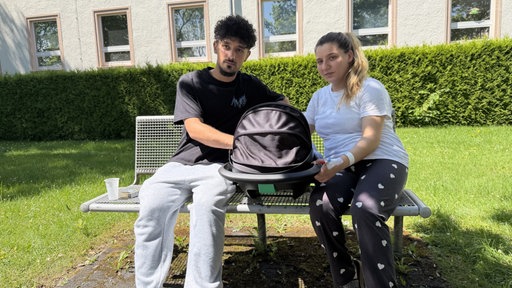 Junge Eltern sitzen mit einer Babyschale auf einer Parkbank.