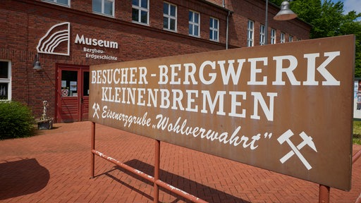 Vor dem Gebäude des Museums steht ein großes Schild mit der Aufschrift „Besucher-Bergwerk Kleinenbremen – Eisenerzgrube Wohlverwahrt“.