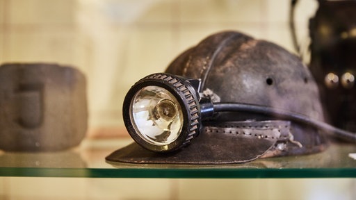 Ein alter Bergmannshelm mit einer daran befestigten Grubenlampe liegt in einer Museumsvitrine.