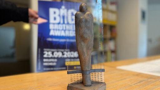 ine dunkle Statuette der Big Brother Awards steht auf einem Holztisch, im Hintergrund ist ein Plakat der Veranstaltung zu sehen.