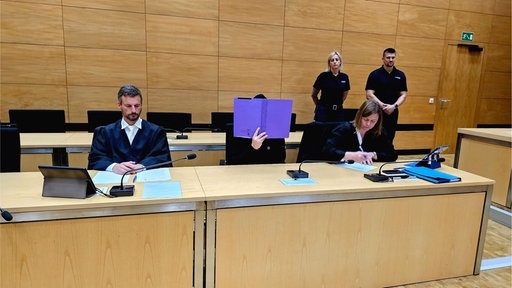Drei Personen sitzen an einem Tisch in einem Gerichtssaal, während zwei weitere Personen im Hintergrund stehen.