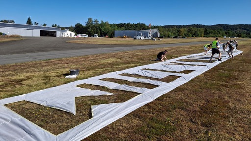 Mehrere Menschen legen auf einer Wiese am Flugplatz eine große Folie mit Buchstaben aus.