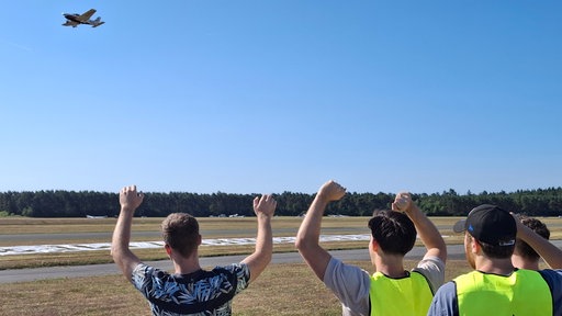 Vier Menschen stehen mit erhobenen Armen am Rand eines Flugplatzes und schauen zur Landebahn.
