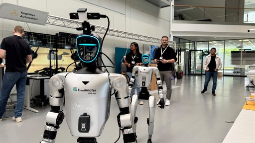 Zwei humanoide Roboter stehen hintereinander in einer Halle, während sich mehrere Personen im Hintergrund befinden.