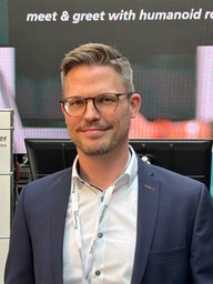 Professor Klaus Neumann steht vor einem Bildschirm und einem Messestand mit Fraunhofer-Schriftzügen.