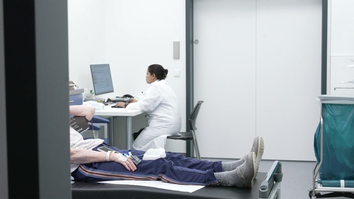 Eine Ärztin arbeitet am Computer, im Vordergrund ein Patient auf einer Behandlungsliege.