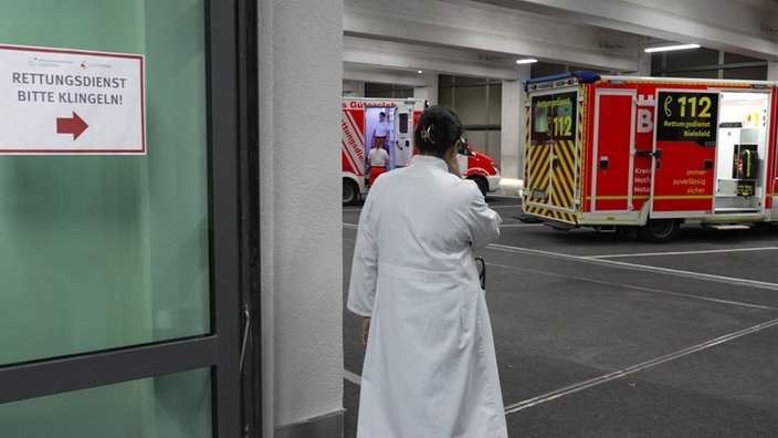 Eine Ärztin im weißen Kittel steht in der Tür zur Rettungszufahrt, davor zwei Krankenwagen.