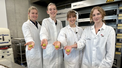 Vier Studentinnen stehen vor einem Blech mit Keksen und halten jeder einen in der Hand. 