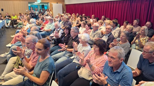 Mehrere Menschen sitzen in einem Saal auf Stühlen und applaudieren.