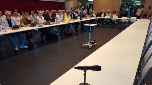 Mehrere Menschen sitzen an U-förmig angeordneten Tischen in einem Saal, während weitere Personen im Hintergrund stehen oder sitzen.