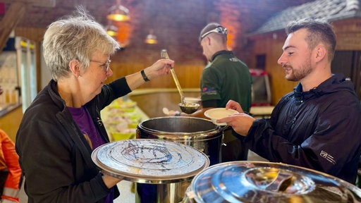 Eine Frau schöpft mit einer Kelle Essen aus einem großen Topf in eine Schale, die ein Mann hält.