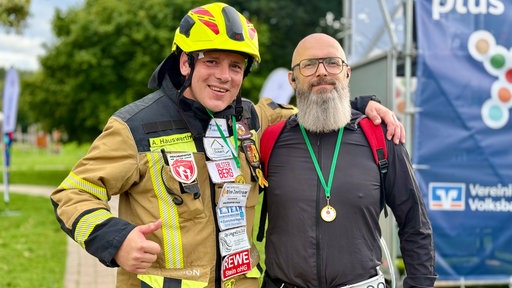 Zwei Teilnehmer stehen nebeneinander, einer in Feuerwehrkleidung, der andere mit einer Medaille um den Hals.