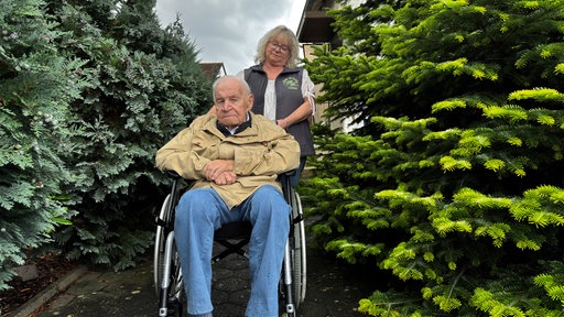 Ein älterer Mann sitzt im Rollstuhl auf einem Gartenweg, während eine Pflegerin hinter ihm steht.