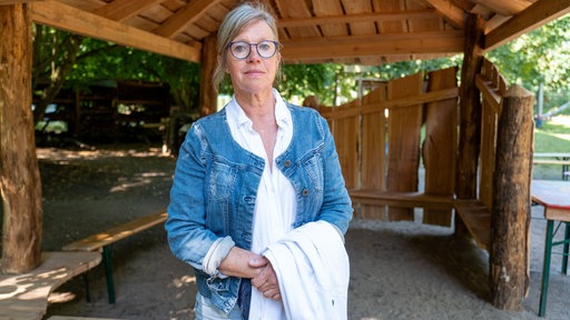 Eine Frau blickt vor einer Holzhütte in die Kamera.