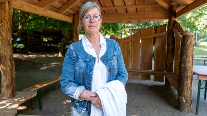 Eine Frau blickt vor einer Holzhütte in die Kamera.