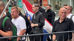 Ein Mann mit Seitenscheitel, Oberlippenbart und in schwarz gekleidet läuft in einer Gruppe mit einer großen niederländischen Flagge in der Hand.