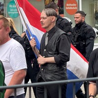 Ein Mann mit Seitenscheitel, Oberlippenbart und in schwarz gekleidet läuft in einer Gruppe mit einer großen niederländischen Flagge in der Hand.