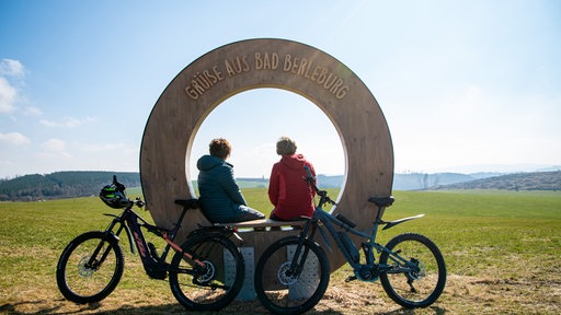 Vor einer Holzskulptur mit der Aufschrift „Grüße aus Bad Berleburg“ stehen zwei Fahrräder, während zwei Personen darin sitzen.