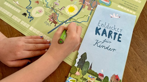 Hände zeichnen mit einem Stift auf einer Karte neben einer Broschüre mit der Aufschrift „Entdecker Karte für Kinder“.