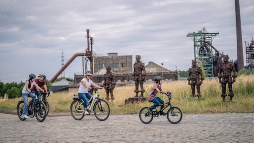 Mehrere Menschen fahren mit Fahrrädern an einer Industrieanlage und Metallskulpturen vorbei.