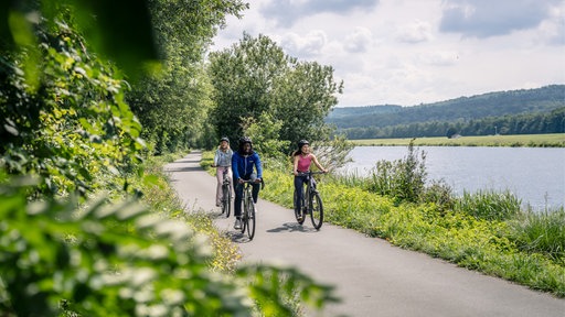 Drei Menschen fahren mit Fahrrädern auf einem Radweg entlang eines Flusses.