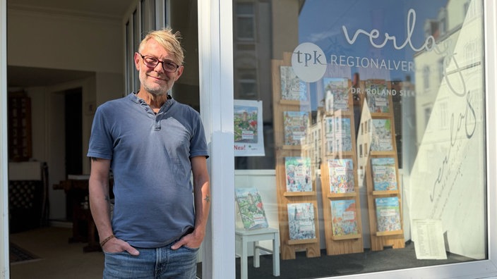 Roland Siekmann steht vor dem Ladengeschäft des tpk Verlags, im Schaufenster stehen mehrere Wimmelbücher.