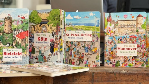 Mehrere Wimmelbücher stehen nebeneinander aufgereiht, darunter die Ausgaben zu Bielefeld, Köln, St. Peter-Ording und Hannover.