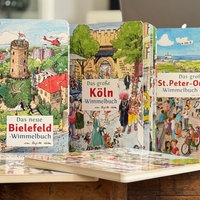 Mehrere Wimmelbücher stehen nebeneinander aufgereiht, darunter die Ausgaben zu Bielefeld, Köln, St. Peter-Ording und Hannover.