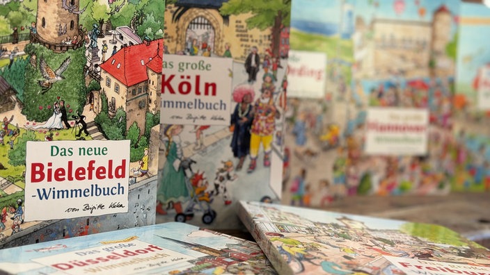 Nahaufnahme mehrerer Wimmelbücher; scharf im Vordergrund die Ausgaben aus Bielefeld und Köln, die übrigen stehen unscharf dahinter.