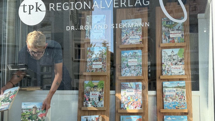 Blick von außen durch das Schaufenster des tpk Verlags auf die ausgestellten Wimmelbücher, links im Bild Roland Siekmann.