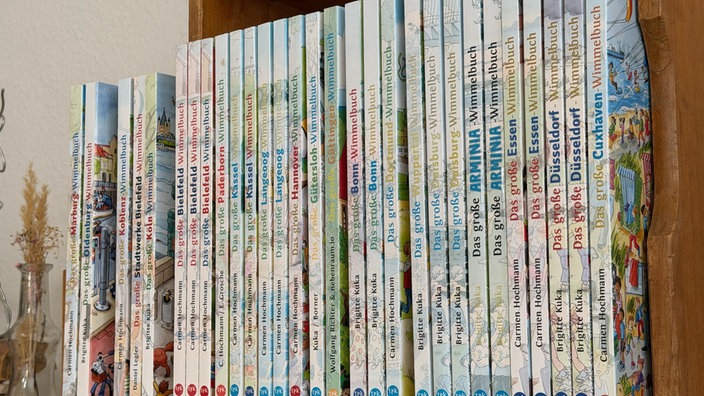 Wimmelbücher aus vielen verschiedenen Städten stehen aufgereiht in einem Regal.