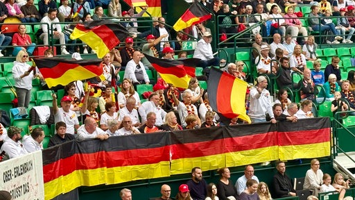 Zahlreiche Zuschauerinnen und Zuschauer sitzen auf einer Tribüne und schwenken Deutschlandflaggen.