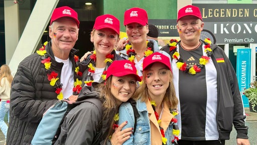 Sechs Tennisfans stehen gemeinsam vor dem Stadion und tragen rote Kappen sowie schwarz-rot-goldene Fanartikel.