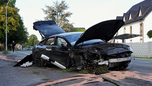 Ein stark beschädigtes schwarzes Auto steht mit geöffneter Motorhaube und Heckklappe am Straßenrand.