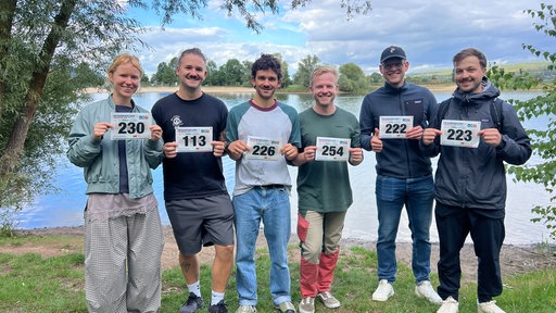 Sechs Personen stehen mit ihren Startnummern vor einem See.
