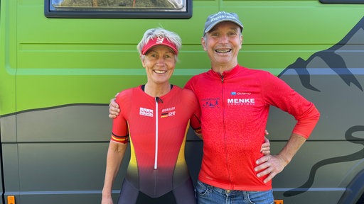Zwei Personen in roter Sportkleidung stehen vor einem grünen Wohnmobil.