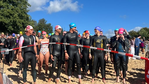 Triathletinnen und Triathleten in Neoprenanzügen warten hinter einer Absperrung am See.