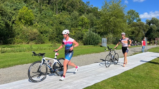 Zwei Triathleten schieben ihre Fahrräder über eine ausgelegte Matte.