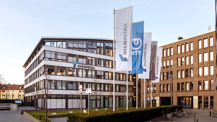 Mehrere Fahnen mit der Aufschrift "Rheinmetall" und Logo stehen vor einem modernen Bürogebäude mit Glasfassade.