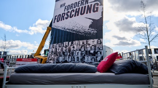 Ein Bett steht vor einem Plakat mit der Aufschrift "Wir fordern Forschung"