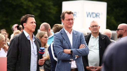 Hendrik Wüst steht in einer Menschenmenge und neben ihm ein Moderator